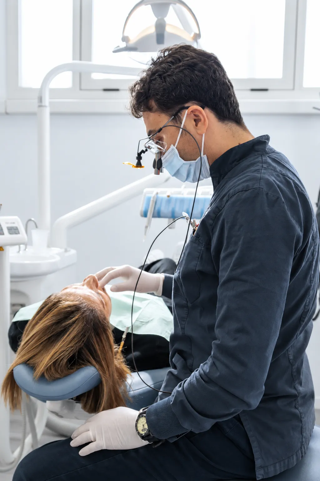 Un dentista in divisa blu scuro, con mascherina e occhiali ingranditori, visita un paziente sdraiato sulla poltrona, in una sala luminosa.