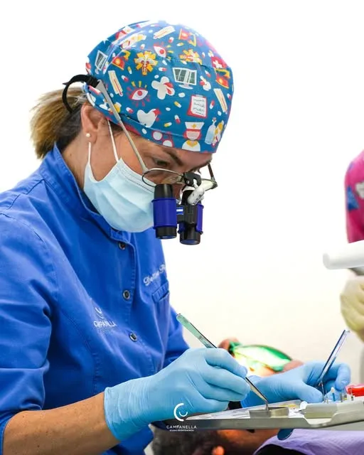 La dottoressa Luisanna, con cuffia a fantasia, occhiali ingranditori e la divisa blu dello studio, al lavoro sul vassoio degli strumenti.